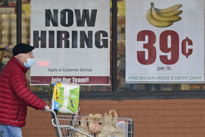 A man with a face mask covering only his mouth pushes a grocery cart outside a store window with a now hiring sign.