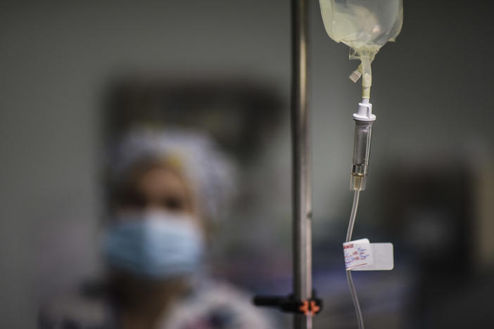 An IV bag hangs in the foreground with a blurred health care worker in the background.