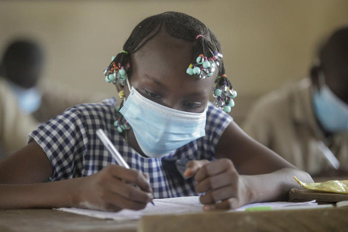 A girl in a mask writes on paper at a school desk. 