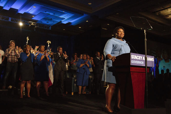 Stacey Abrams stands on stage with supporters clapping behind here.