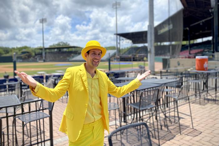 Savannah Bananas founder and owner Jesse Cole poses in a patio area at an empty Grayson Stadium. He is wearing a yellow tuxedo and yellow top hat.