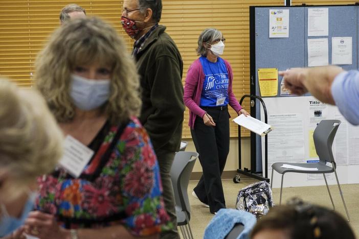 A scene from an election polling site shows people waiting in line and a poll watcher carrying a clipboard.