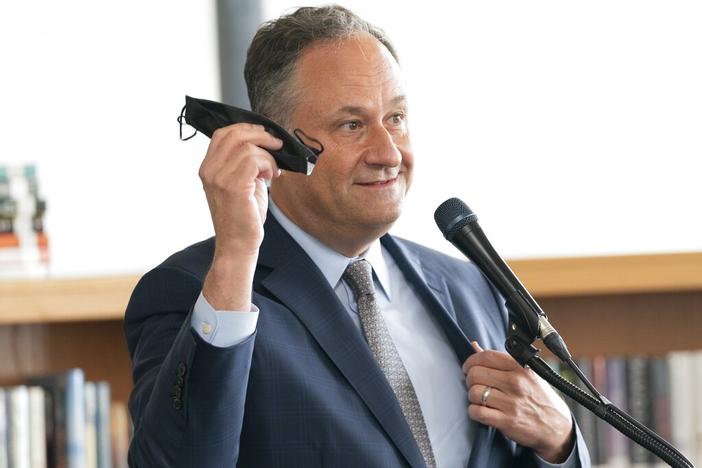 Douglas Emhoff holds a Covid mask in his hand while speaking behind a microphone.