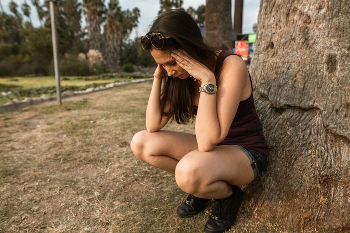 A woman kneels in front of a tree with her hands on her temples