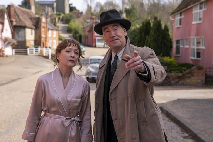 A woman in a robe and a man in a hat and coat looking down a sunny street.