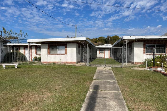 Squat one-story buildings surrounded by chain link fence comprise a former motel.