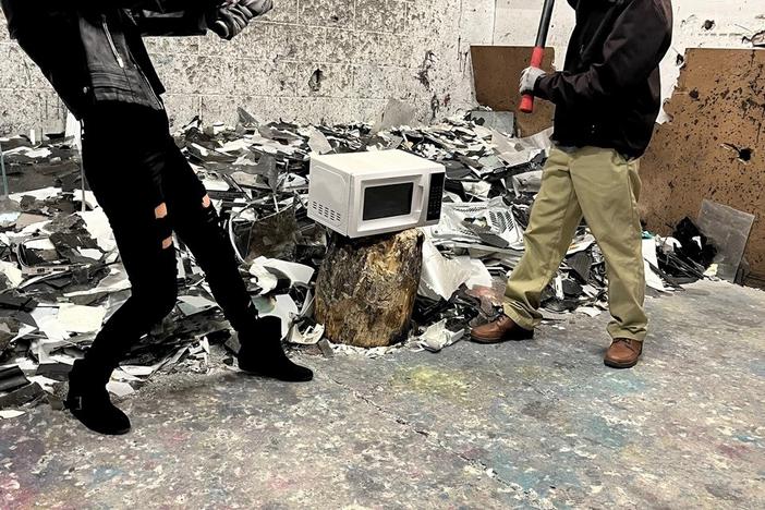 Two people, one wielding a bat and the other a sledgehammer, are poised to smash a small microwave in a cinderblock room.