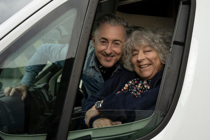 Miriam Margolyes and Alan Cumming smiling in the window of a mobile home.