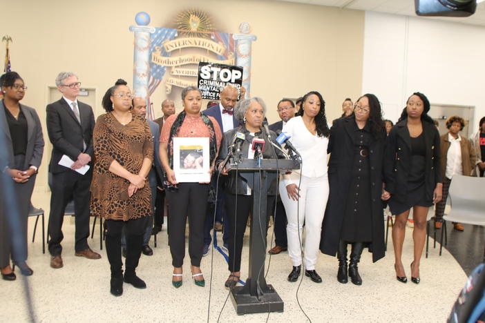 Chris Ann Lewis is shown speaking behind a podium into microphones while her family members stand behind her.