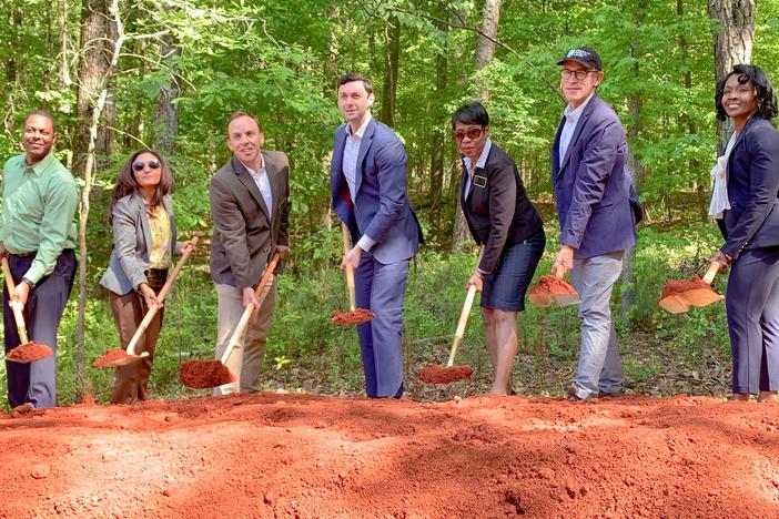 Seven people are shown holding shovels in a groundbreaking ceremony in a wooded environment.