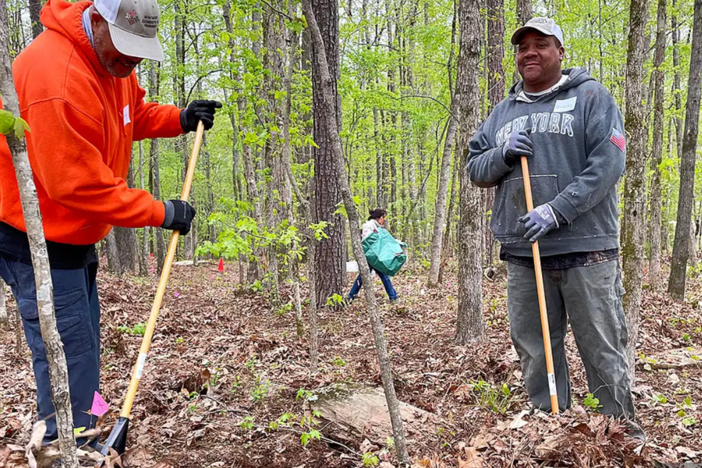 Two men hold rakes in a forested scene.