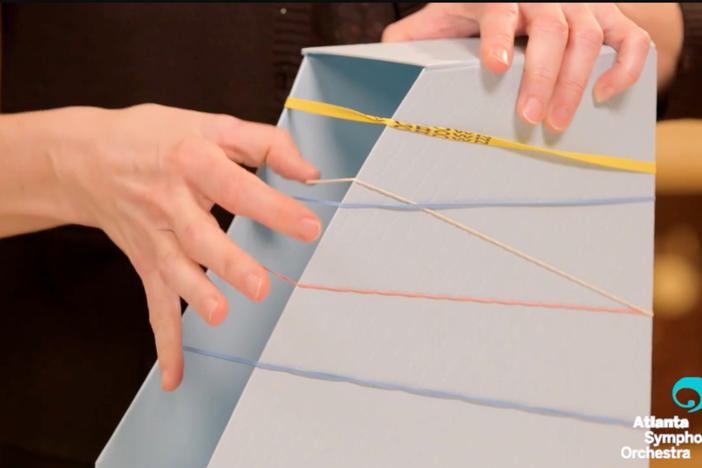 Musician stretches rubber band around an open filing box to create an instrument.