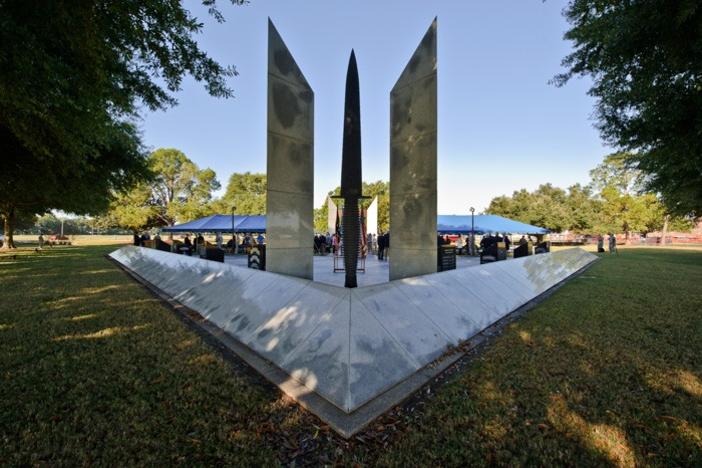 A stone monument is shown in a grassy, tree-lined field.