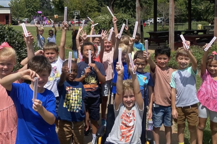 A class of young students stands outside holding their science experiments up for the camera with pride.