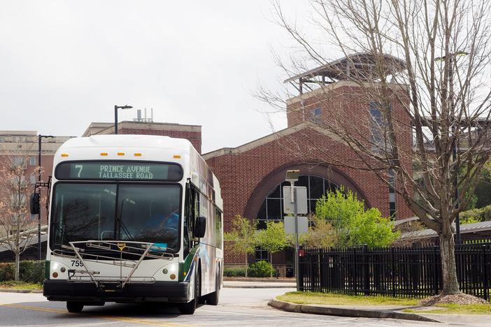A bus is shown, pictured from the front, driving out of a driveway with a red brick building behind it.