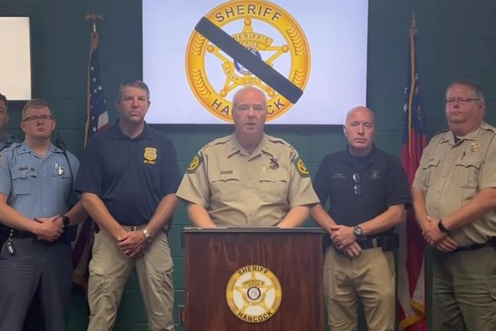 Crisp County Sheriff Billy Hancock is shown behind a podium with law enforcement members standing by his side.