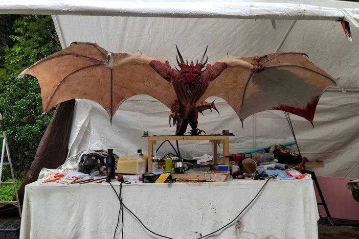 A mock-up of a horned, winged dragon sits on a work table with tools underneath a canopy.