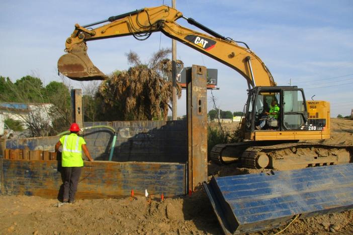 A backhoe is used at a construction site in Savannah, with a construction worker watching from a distance.