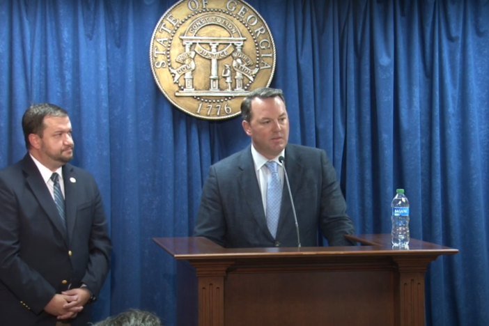 Burt Jones is seen speaking at a podium in front of a plain curtain backdrop and an official Georgia seal.  Dallas State Senator Jason Anavitarte is shown standing to his left.