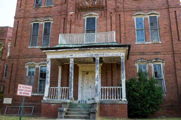 The run-down facade of an old brick building is shown with a sign in front of it that says "No Trespassing Unsafe Building and Grounds."