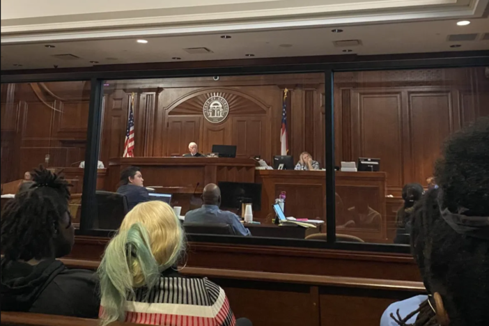 Public defender Tobe Karrh (left in suit) looks on at evidence projected onto a screen in the felony murder trial of his client Varshan Brown (center in blue shirt) on Sept. 12, 2023, in the killing of his cousin Latoya James. James was killed by police during a drug raid, according to court testimony.