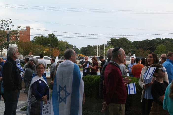 A group of people, some carrying Israeli flags, is shown standing outside.