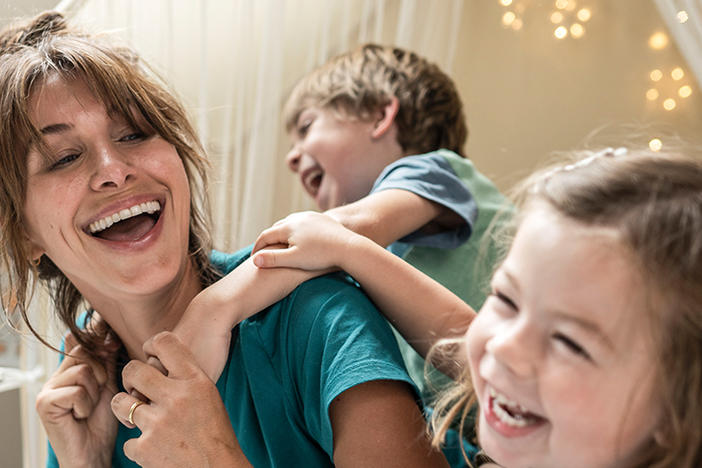 A woman in scrubs with a young boy on her back and a young girl is smiling