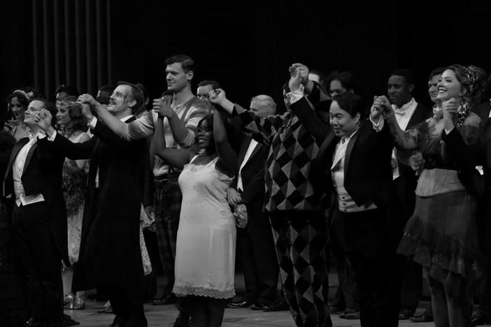 A Black and white photo shows the opera performers holding hands at the edge of the state preparing to take a bow.