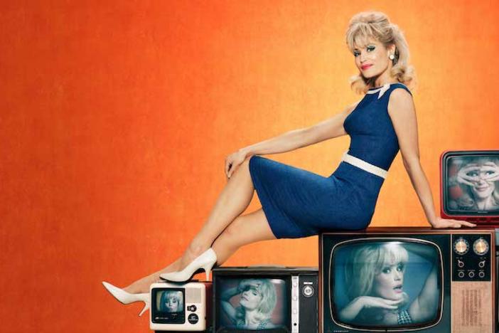 A blonde woman in 60s period dress sitting on top of old televisions.