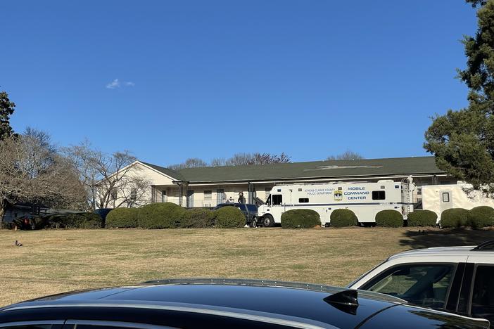A police mobile command center vehicle is shown in the distance across a grass lawn and in front of apartment unites.