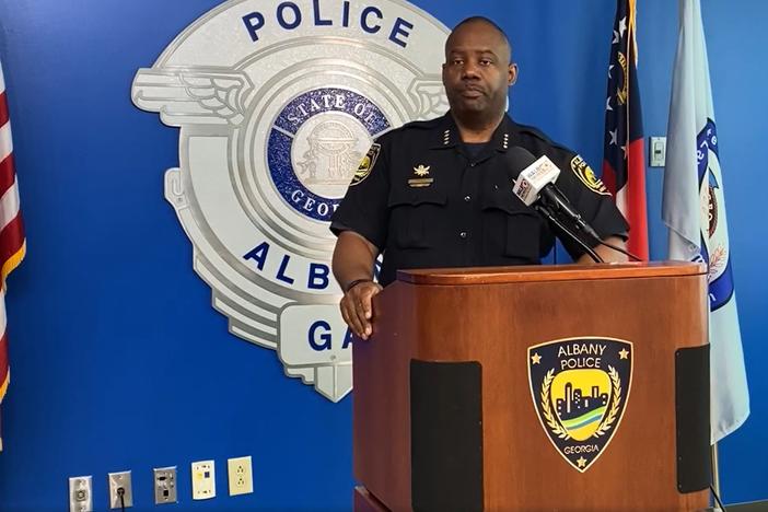 Albany Police Chief Michael Persley is shown standing at a podium, indoors and flanked by three flags.
