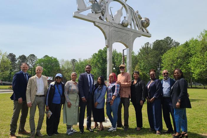 the mayor and a group of artists stand shoulder to shoulder in the grass in front of a large metal monument. It has an arch with a flat top where music objects, a globe, and the face of Martin Luther King are visible on top.