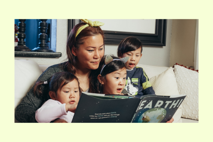 A grownup sits with three kids as they all look at a book. 