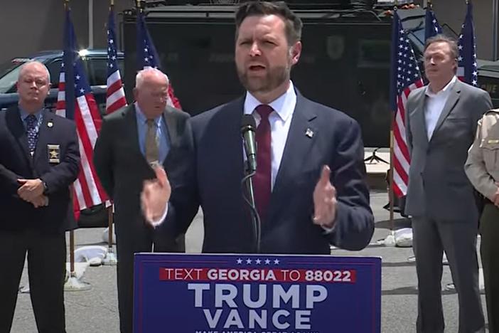 Ohio Senator JD Vance is show speaking from behind a podium.  He is flanked by other men and flags.