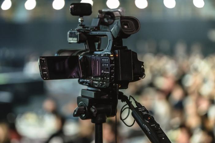 lose-up of a video camera mounted on a tripod, with a blurred audience in the background.