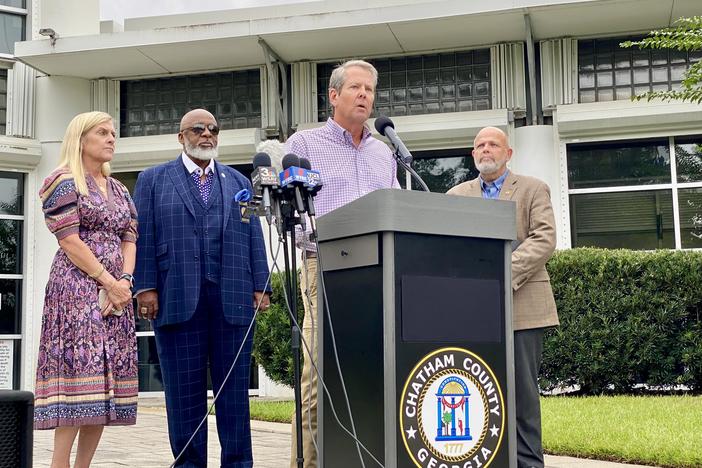 Governor Brian Kemp is shown speaking behind a podium, surrounded by officials from Chatham County.