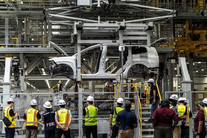 The unfinished frame of a sports utility vehicle hangs above a group of about a dozen people wearing safety vests and hard hats inside a factory.