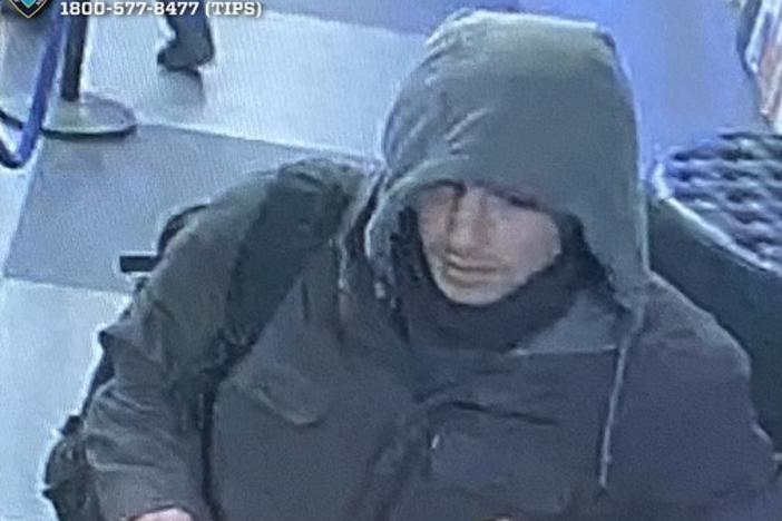 A grainy surveillance video image shows a person wearing heavy grey coat.  The person's face is partially obscured by the coat's hood.