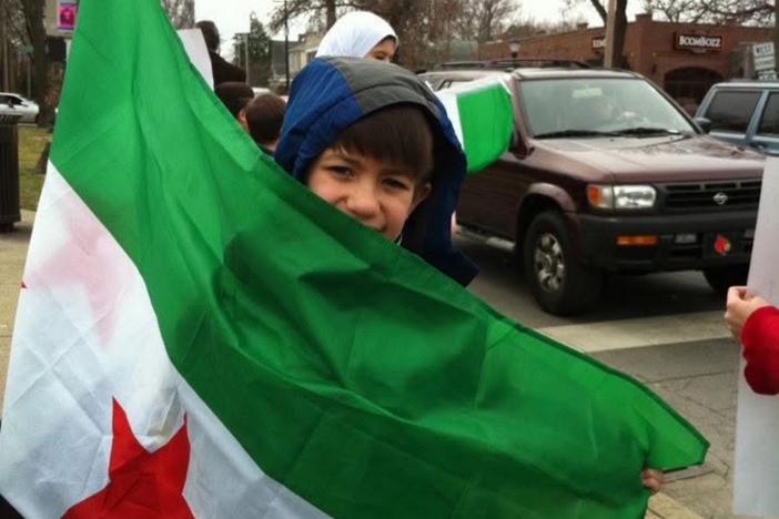 A child is shown in a parking lot holding a very large Syrian flag.
