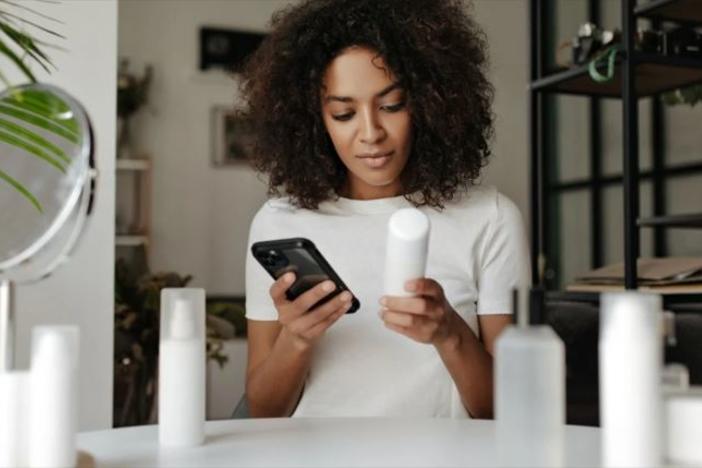 A Black woman looks at a beauty product and holds a cellphone