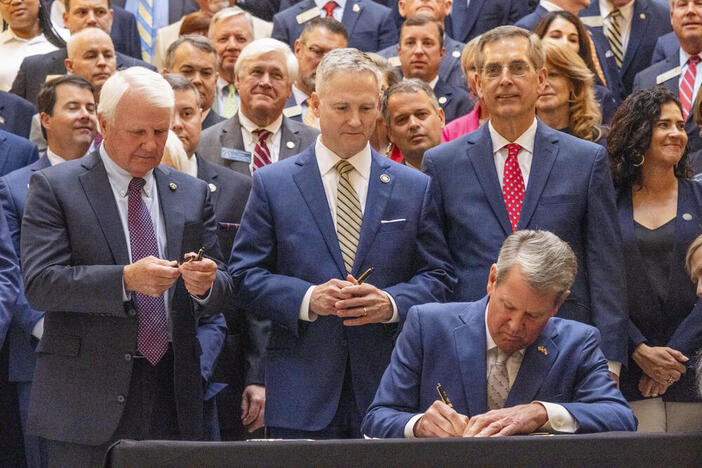 Governor Brian Kemp signs a bill on a desk, with dozens of supporters standing behind him.