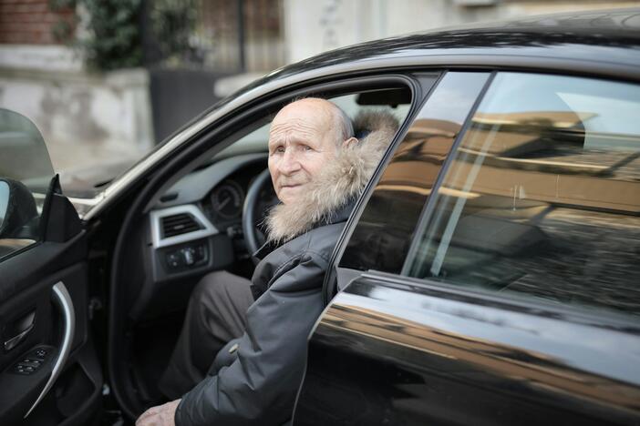 A man in the driver's seat of a car wearing a black jacket and looking out of the open car door