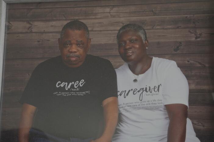 An older black man and woman sitting next to each other. The man has a black shirt that says caree and the woman has a white shirt that says caregiver.