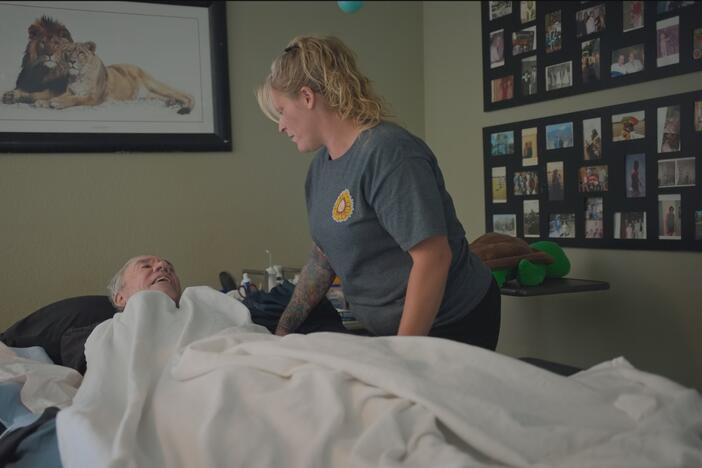 Woman with a blond ponytail speaking with an older man lying on a bed covered in a white blanket