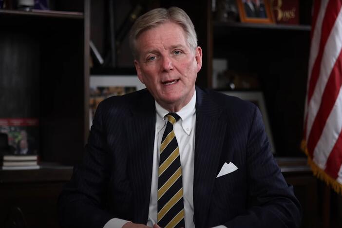 Brant Frost IV is shown seated and speaking to the camera in his office with an American flag in the background.