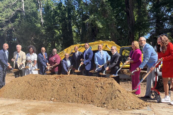 Twelve people holding shovels like they're going to dig stand in suits behind a pile of dirt.