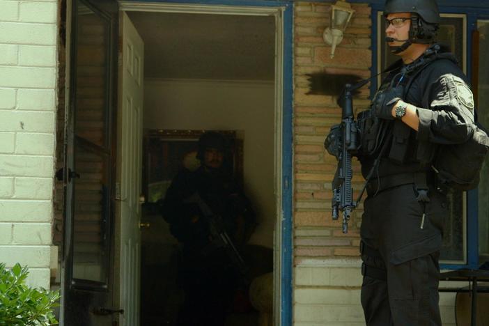The clip shows a military-equipped police SWAT team conducting a drug raid.