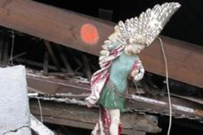 In the ruins of a collapsed church, a statue of an angel, missing a wing but upright.