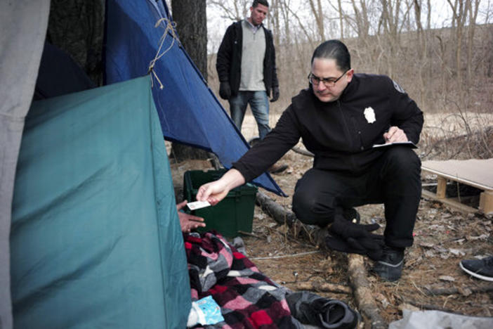 Officer Angel Rivera, right, returns a license to an unidentified man as Rivera asks if he has been tested for Hepatitis A at the entrance to a tent where the man spent the night in a wooded area in Worcester, Mass, after an outbreak of hepatitis A.