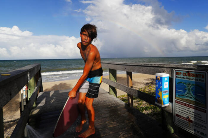 A boy cleans his surfboard as a rainbow is seen over the Atlantic Ocean along the beach in Vero Beach, Fla., Saturday, Aug. 31, 2019.